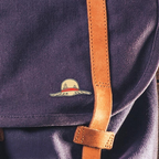 Fans Geek Store metal pin featuring Luffy's iconic straw hat from One Piece attached to a purple canvas backpack