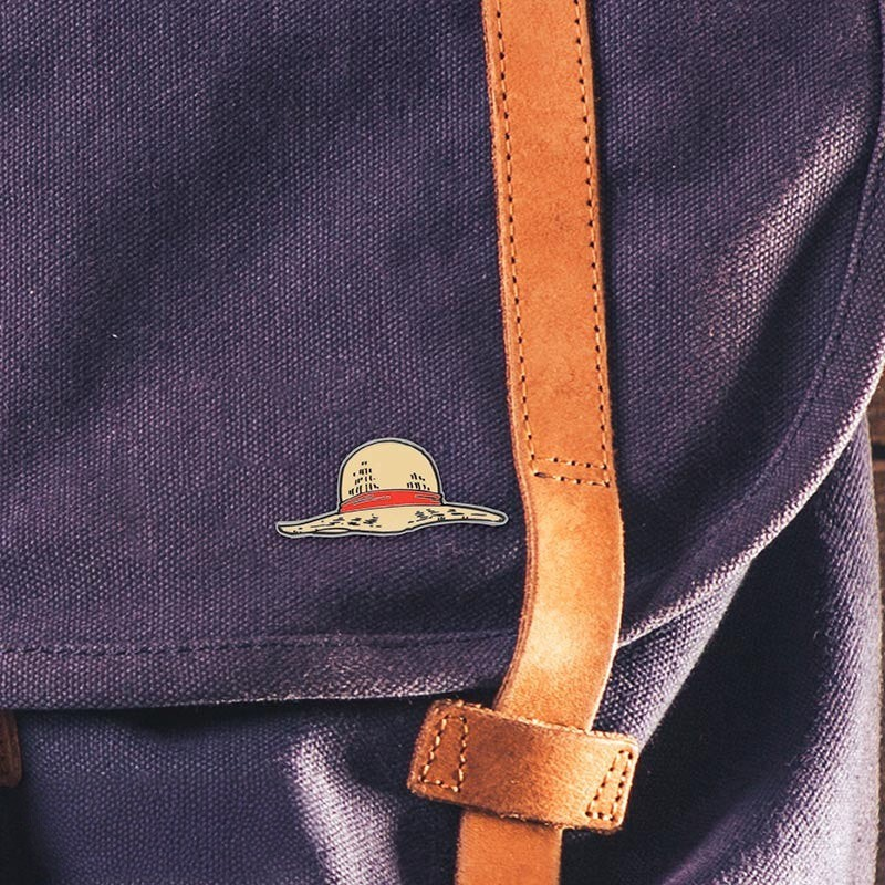 Fans Geek Store metal pin featuring Luffy's iconic straw hat from One Piece attached to a purple canvas backpack